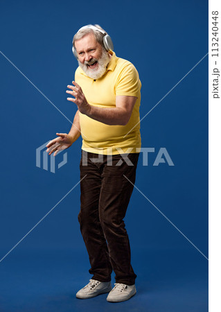 Elderly bearded man in yellow polo listening to music in headphones and dancing against blue studio background. Full-length 113240448