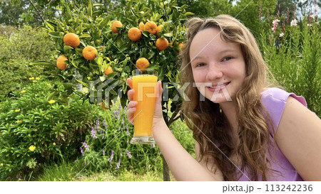 a teenage girl stands with a glass of orange juice on the background of a citrus tree tangerines or oranges hang on a tree. She smiles and looks into the frame can be used to advertise juices a teenage girl stands with a glass of orange juice on the background of a citrus tree tangerines or oranges hang on a tree. She smiles and looks into the frame can be used to advertise juices 113242236