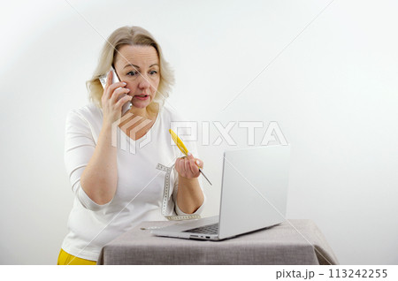 serious dissatisfied woman talking on the phone looks into a laptop in her hands scissors and a meter weight loss diet tired of communicating with a doctor nutritionist middle age white background serious dissatisfied woman talking on the phone looks into a laptop in her hands scissors and a meter weight loss diet tired of communicating with a doctor nutritionist middle age white background 113242255