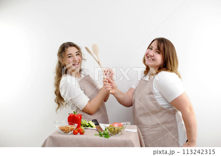 two women two friends fight on wooden spoons smile laugh in the frame play have fun on the table knife vegetables nuts aprons of the same color on a white background cooking cooking grocery store 113242315