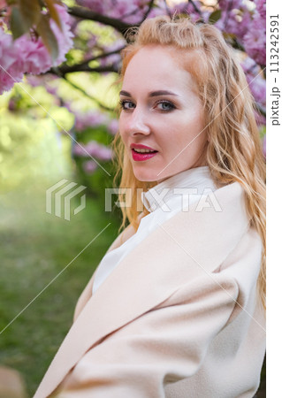 Red-haired girl with pink lipstick stands near pink flowering tree looks into the frame and smiles. Attractive young woman. Vertical frame. 113242591