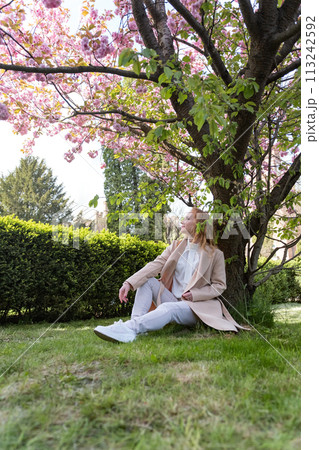 Young woman sitting on lawn in blossoming park, warm spring day. Attractive girl on rest. Vertical frame. Young woman sitting on lawn in blossoming park, warm spring day. Attractive girl on rest. Vertical frame. 113242592