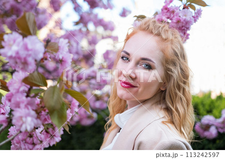 Portrait of happy attractive woman in beige coat among blossoming sakura branches. Stylish girl with red lips in blooming park. 113242597