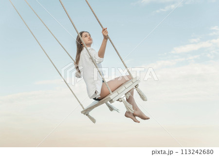 Young beautiful girl in white shirt on rope swing high on blue sky background during a sunset time. Young beautiful girl in white shirt on rope swing high on blue sky background during a sunset time. 113242680