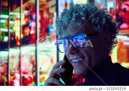 Smiling curly man standing near big city mall in eyewear with urban lights reflection in glasses and talking on mobile phone. Evening city scene Smiling curly man standing near big city mall in eyewear with urban lights reflection in glasses and talking on mobile phone. Evening city scene 113243314