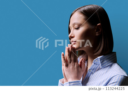 Close-up profile view of young woman with folded hands on blue background 113244225