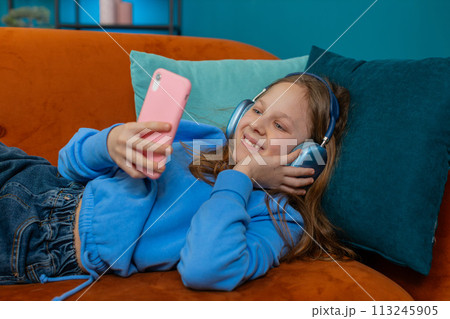 Happy Caucasian girl child in wireless headphones relaxing lying on couch at home listening music Happy Caucasian girl child in wireless headphones relaxing lying on couch at home listening music 113245905
