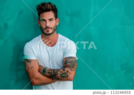 A man with tattoos on his arms stands in front of a green wall 113246958