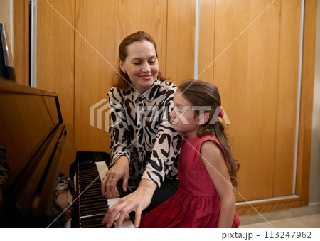 Cheerful mother and daughter pianists playing piano together, performing a melody for Christmas during music lesson together. Cute baby girl in elegant red dress, listens to her mom playing pianoforte 113247962