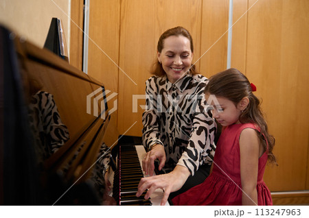 Cheerful beautiful woman pianist, musician teacher smiling while performing melody on piano, explaining music lesson to her student girl 113247963