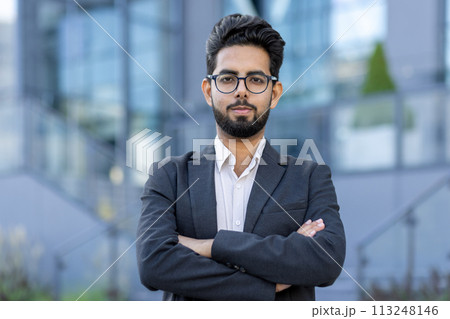Portrait of a smiling and successful young Indian businessman in a suit standing outside on the street, crossing his arms over his chest and looking confidently at the camera. Portrait of a smiling and successful young Indian businessman in a suit standing outside on the street, crossing his arms over his chest and looking confidently at the camera. 113248146