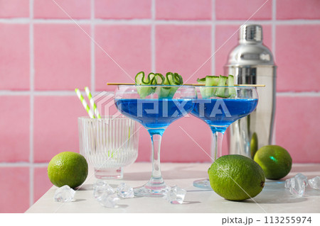 Blue cocktail with cucumber slice in glasses, shaker, limes and ice cubes on pink background, close up 113255974