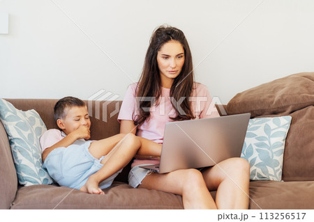 Young mother trying to work on laptop at home while her son wants to play. Childcare and working mom concept. Child make noise and disturb a woman at work. Homeschooling and freelance work. Young mother trying to work on laptop at home while her son wants to play. Childcare and working mom concept. Child make noise and disturb a woman at work. Homeschooling and freelance work. 113256517