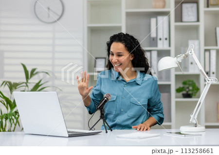 Cheerful professional woman waving and smiling during a podcast recording in a bright home office setup. Cheerful professional woman waving and smiling during a podcast recording in a bright home office setup. 113258661