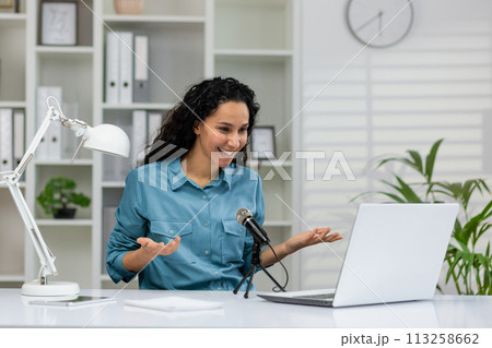 Cheerful professional woman recording a podcast in a contemporary home office set up with a laptop and microphone. Cheerful professional woman recording a podcast in a contemporary home office set up with a laptop and microphone. 113258662