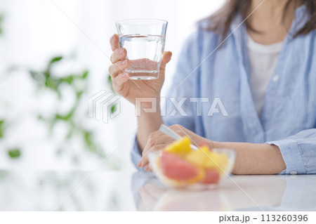 水のグラスを手にフルーツを食べるミドル女性の手元 113260396