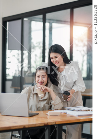 Two smiling female colleagues collaboratively working on a project with a laptop and coffee in a bright office space. 113261529