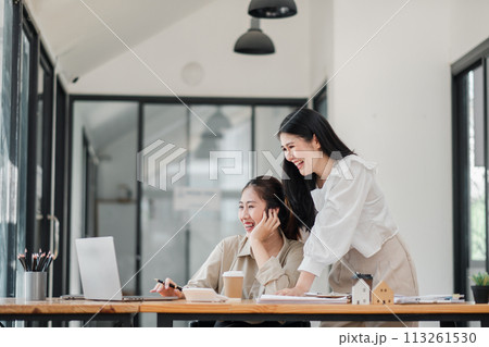 Two joyful colleagues share a light-hearted moment, with one seated at a laptop and the other standing beside her, in a spacious and well-lit office. 113261530