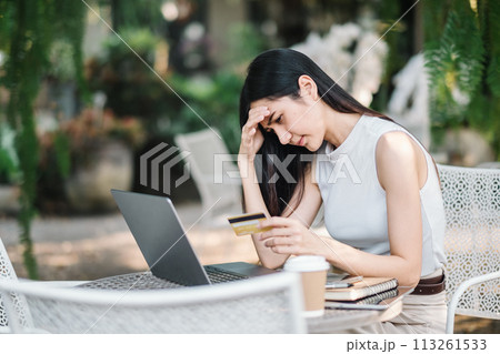 Worried young woman experiencing problems with an online payment, holding a credit card and looking at laptop screen at an outdoor cafe. 113261533