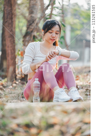 Smiling woman in sportswear checking her fitness tracker after exercising, seated in a serene forest area. Smiling woman in sportswear checking her fitness tracker after exercising, seated in a serene forest area. 113261585