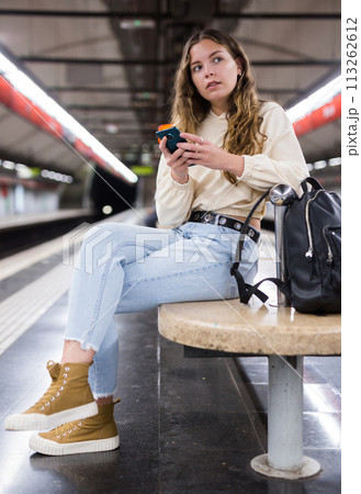 Portrait of a pensive girl sitting on a subway platform bench with a mobile phone 113262612