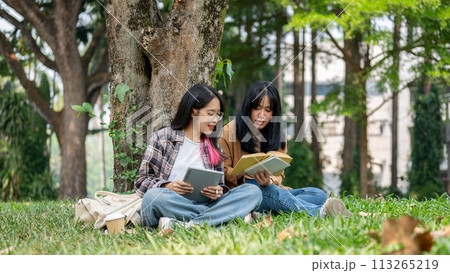 Two young Asian female students sit on the grass under a tree, studying together in a campus park. 113265219