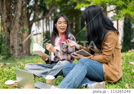 Two young Asian female students are sitting on the grass in a park, studying together. 113265244