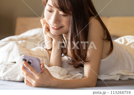 ベッドに寝転んでスマートフォンを使う笑顔の女性 ベッドに寝転んでスマートフォンを使う笑顔の女性 113268759