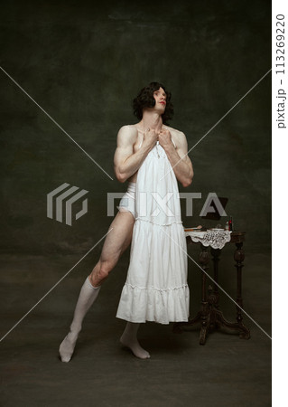 Athletic man fitting underdress showing combination of masculinity and traditional femininity against vintage studio backdrop. Athletic man fitting underdress showing combination of masculinity and traditional femininity against vintage studio backdrop. 113269220