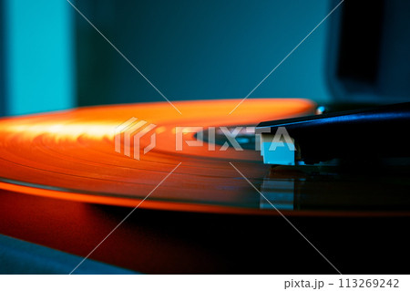 Detailed photo of phonograph needle riding waves of vinyl's surface in orange hues. Retro vinyl record player, bright lights. 113269242