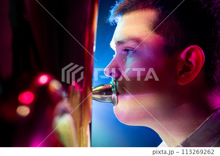 Close up side view photo of talented man, artistic musician seated, playing trumpet with soft blue-pink stage lights. 113269262