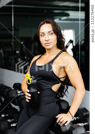 Young woman standing and resting after workout or exercise in fitness gym. Woman at gym taking a break and relax with water in sportswear. Fitness and healthy style concept Young woman standing and resting after workout or exercise in fitness gym. Woman at gym taking a break and relax with water in sportswear. Fitness and healthy style concept 113276466