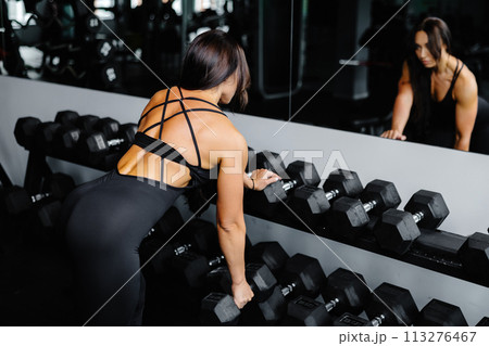 Beautiful woman in sportswear looking at mirror while leaning on rack and lifting heavy dumbbell in gym Beautiful woman in sportswear looking at mirror while leaning on rack and lifting heavy dumbbell in gym 113276467
