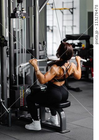 Muscular athletic female bodybuilder in black suit pulled on sports simulator in gym. Back muscles are very tense. Muscular athletic female bodybuilder in black suit pulled on sports simulator in gym. Back muscles are very tense. 113276478