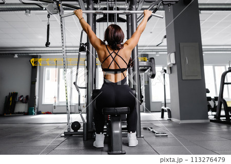 Muscular athletic female bodybuilder in black suit pulled on sports simulator in gym. Back muscles are very tense. Muscular athletic female bodybuilder in black suit pulled on sports simulator in gym. Back muscles are very tense. 113276479