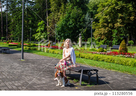Woman and dog - Cavalier King Charles Spaniel - relaxing in a public park with lush flowers, plants, grass and trees 113280556