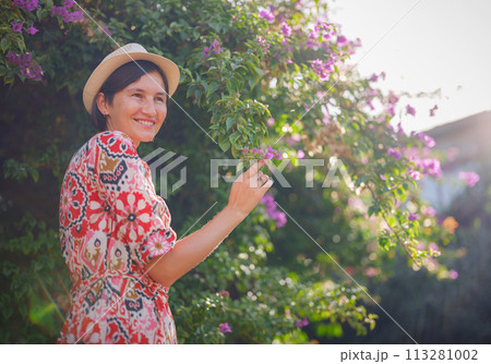 young asian woman stands in sunny green park, admiring blooming bougainvillea bush. The flower's bright colors are beautiful contrast to park's lush greenery. 113281002