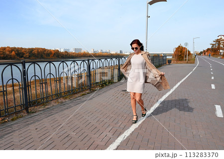 a confident young attractive woman in a coat and high-heeled shoes walks through the streets of the city, the woman smiles and enjoys life 113283370
