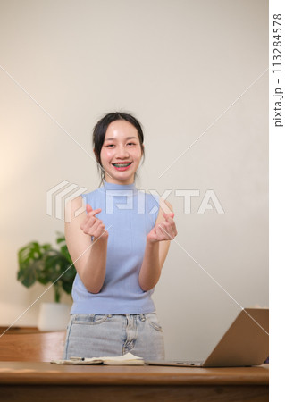 Young attractive happy Asian female student standing in the living room smiling and looking at the camera while working on a laptop at the home office. Young attractive happy Asian female student standing in the living room smiling and looking at the camera while working on a laptop at the home office. 113284578