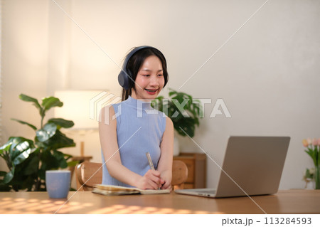 A young female student is sitting at the desk in the workspace and jotting down notes while studying online using a laptop and wearing headphones. 113284593