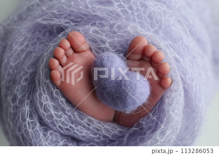 The tiny foot of a newborn baby. Soft feet of a new born in a lilac, purple wool blanket. Close up of toes, heels and feet of a newborn. Knitted purple heart in the legs of a baby. Macro photography.  113286053