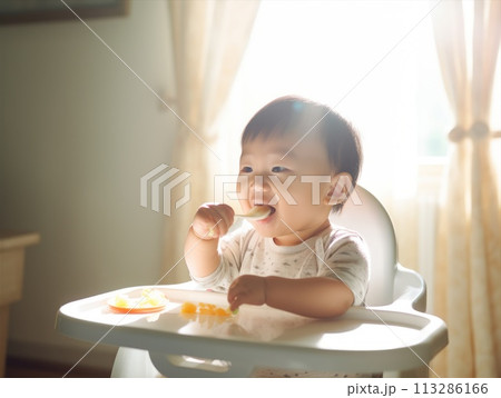 A baby smiling at the table, grabbing food with a spoon from a bowl A baby smiling at the table, grabbing food with a spoon from a bowl 113286166