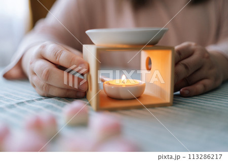 Close-up hands of unrecognizable young woman lighting handmade candle with match sitting at table. Process of making handmade natural scented candle. Creative occupation of making DIY candles. Close-up hands of unrecognizable young woman lighting handmade candle with match sitting at table. Process of making handmade natural scented candle. Creative occupation of making DIY candles. 113286217