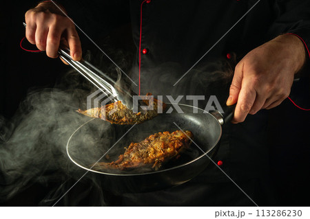 The chef is frying fish in the kitchen. The concept of preparing a fish dish for lunch. The cook hand takes out hot fried crucian carp from the frying pan with tongs 113286230