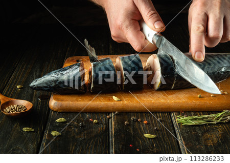 The chef cuts a Scomber fish with a knife on a kitchen cutting board. Menu or recipe for restaurant or hotel. Cook hands close up. Advertising space 113286233