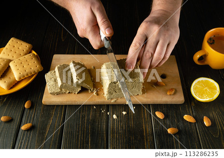 Sliced sunflower halva for dessert. A knife in the hand of a chef for cutting halva for tea with cookies and almond nuts Sliced sunflower halva for dessert. A knife in the hand of a chef for cutting halva for tea with cookies and almond nuts 113286235