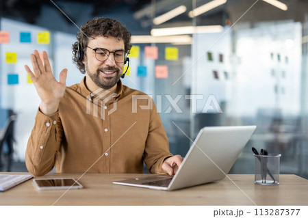 Cheerful businessman having a video call, waving hello to the webcam on his laptop in a modern office setting. 113287377