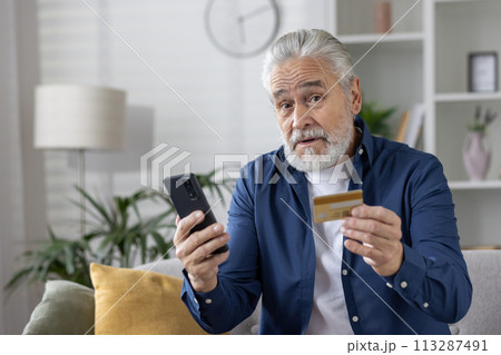 Elderly male with credit card and phone, potential victim of online fraud in a cozy home setting. Elderly male with credit card and phone, potential victim of online fraud in a cozy home setting. 113287491