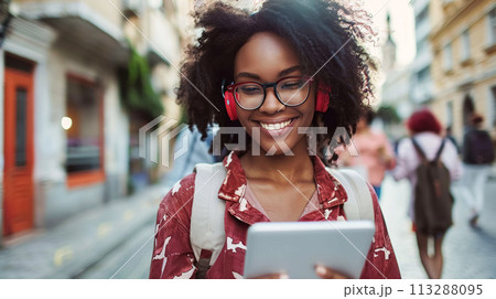 Happy young African woman wearing headphones walks along busy city street and communicates via video call. Modern communication, lifestyle Happy young African woman wearing headphones walks along busy city street and communicates via video call. Modern communication, lifestyle 113288095