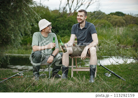Father and adult son with fishing rods near the lake fishing at summer. Fishermen sitting on wooden chair and drinking a beer. Men wearing khaki clothes and a caps. Father and adult son with fishing rods near the lake fishing at summer. Fishermen sitting on wooden chair and drinking a beer. Men wearing khaki clothes and a caps. 113292159
